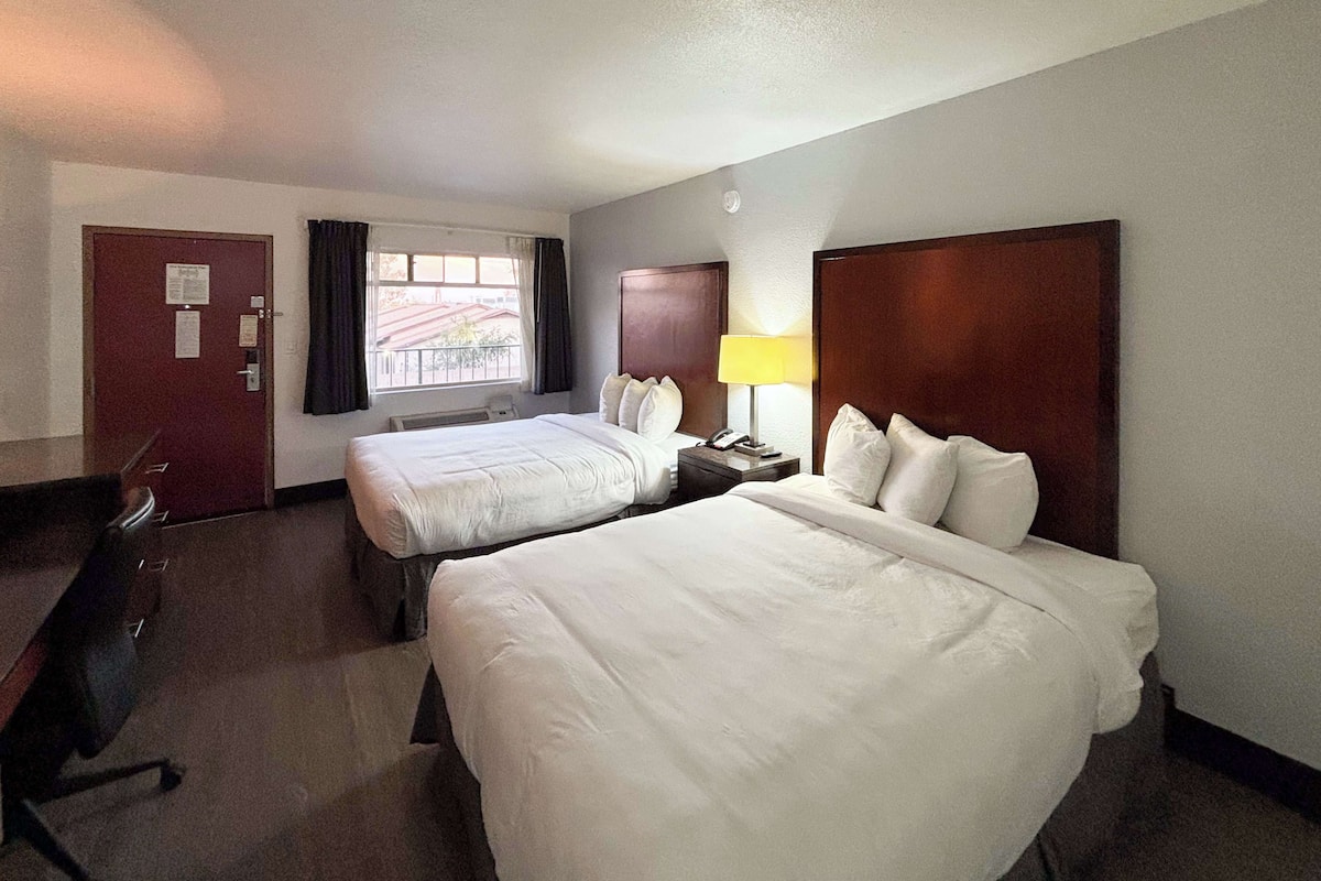 A hotel room with two beds, a desk, a lamp, and a window with curtains.