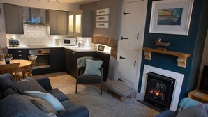 A modern kitchen with grey cabinets, a white countertop, and a built-in microwave. There is a dining table with chairs and a small bench in the living area. - Brakestone Cottage in the heart of Port Isaac (Port Isaac)