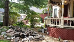 Un jardin avec une petite cascade, une terrasse en bois et un bâtiment rouge en arrière-plan.