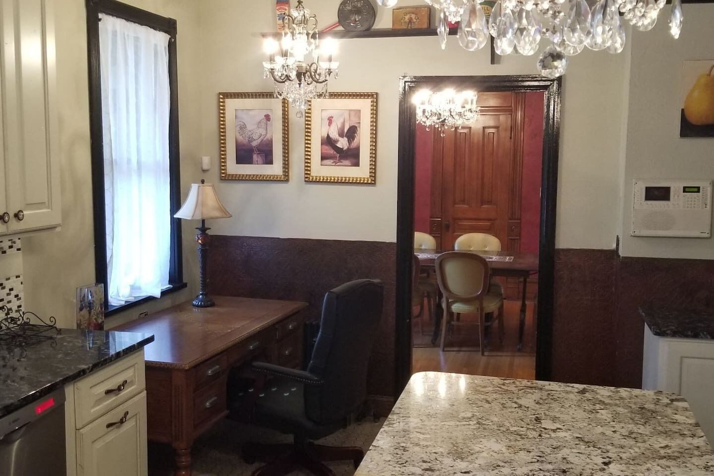 A kitchen with a granite countertop, a wooden desk, a chair, a chandelier, and framed pictures on the wall.