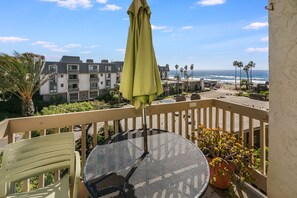 A balcony with a round table and chairs, a yellow umbrella, and a view of a beach and buildings in the background.
