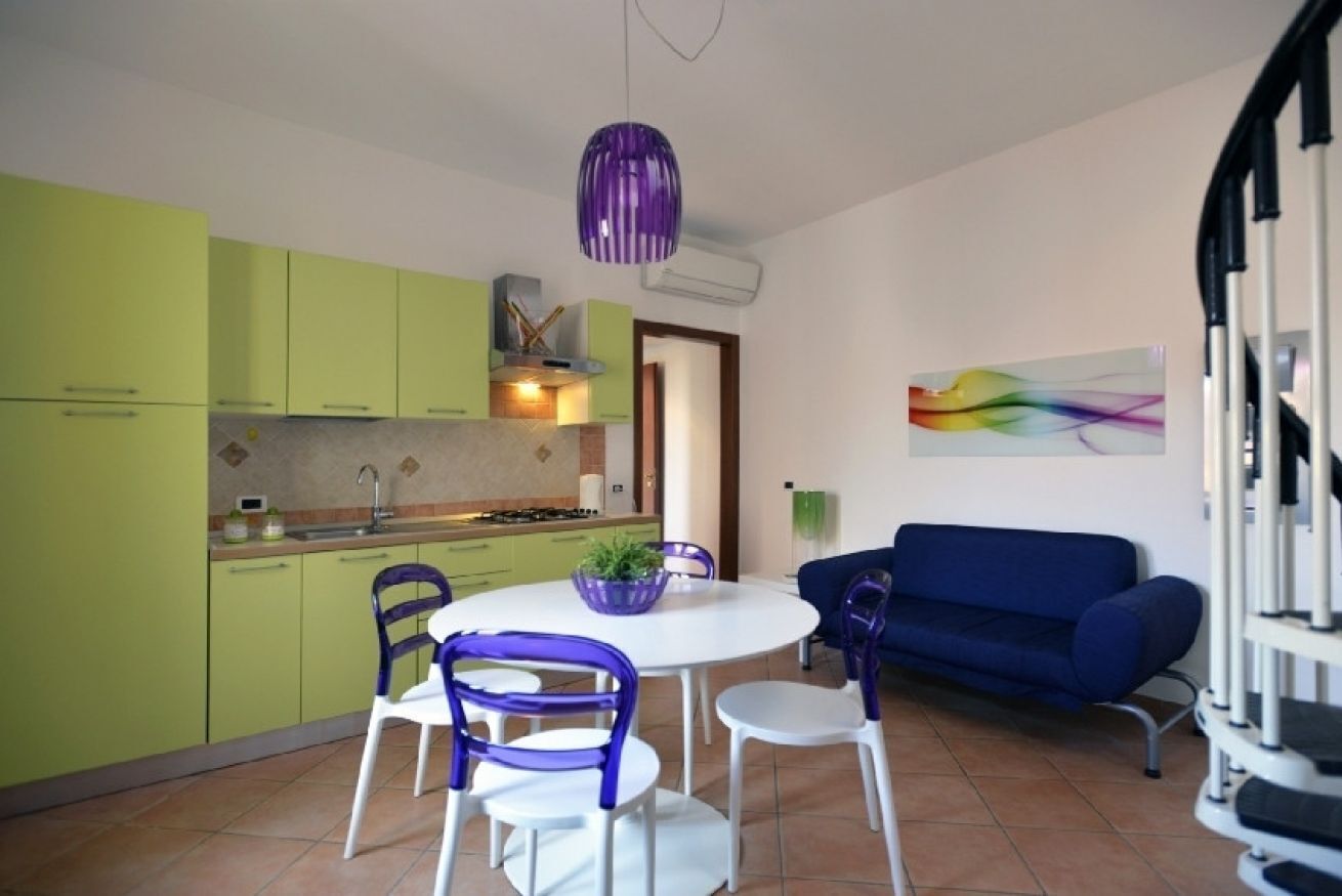 Eine moderne Küche mit grünen Schränken, einem weißen Esstisch mit violetten Stühlen und einem blauen Sofa in einem Wohnbereich.