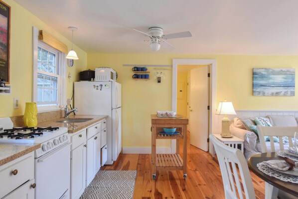 A compact kitchen with white cabinets, a stove, and a refrigerator. There is a wooden table with a bowl and a lamp. A ceiling fan is visible above. - Cottage w/ Private Association Pool! (Wellfleet)