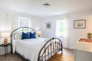 Une chambre à coucher avec un lit en métal, une literie blanche, deux lampes de chevet, une petite table avec une lampe et une fenêtre avec des rideaux.