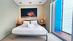 A neatly made bed with white linens, two bedside tables with lamps, a painting above the bed, and a large window with a view. - Beachside living at its best! Bondi Beach Apartment. (Bondi Beach)