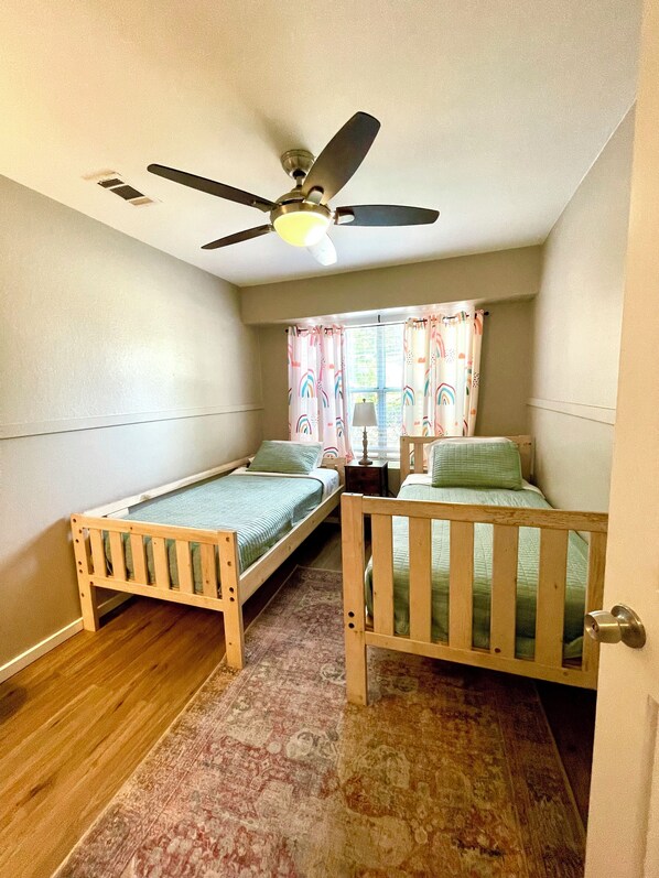 A small room with two wooden beds, a ceiling fan, a window with curtains, and a patterned rug.