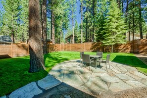 Relax in a serene, shaded backyard patio area with a stone patio, lawn, and tall trees, perfect for outdoor dining and casual gatherings.
