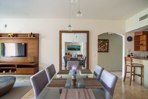 Una sala de estar moderna con una mesa de comedor de vidrio, asientos cómodos, un televisor de pantalla plana y una zona de cocina con armarios de madera.