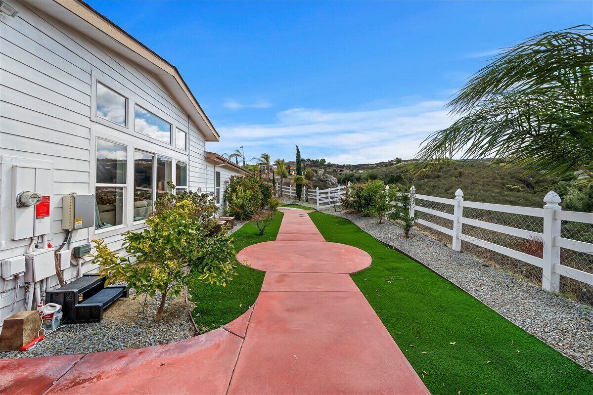 A pathway leading to a white house with a fenced yard and a palm tree.