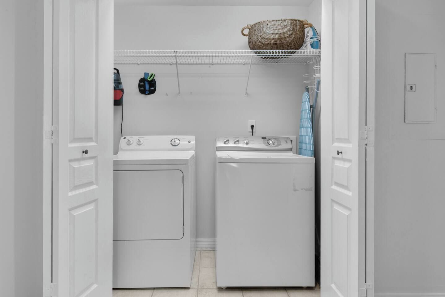 A laundry room with two white washing machines, a wicker basket on the shelf, and a blue towel hanging on the wall.
