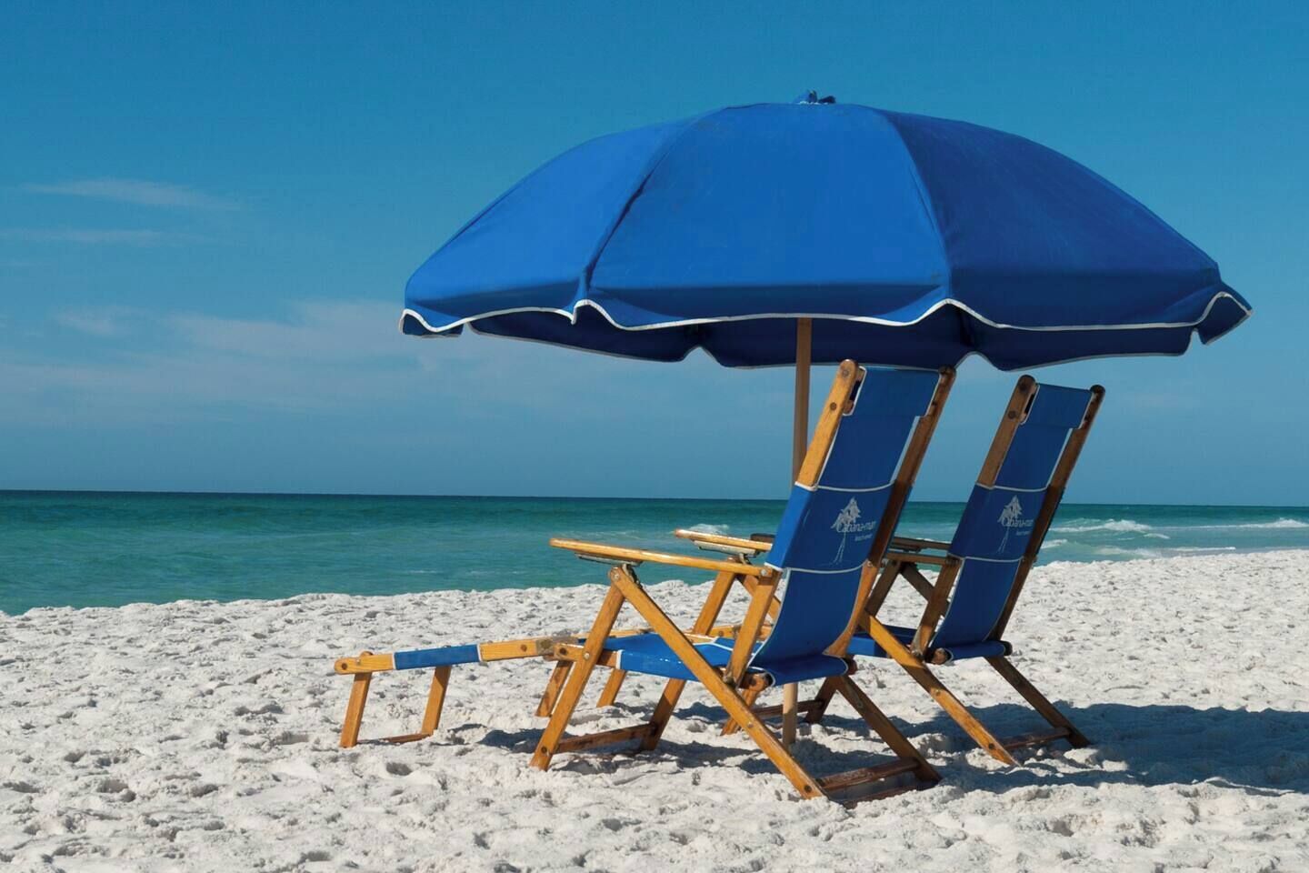 Una playa con un par de sillas de madera y una sombrilla azul.