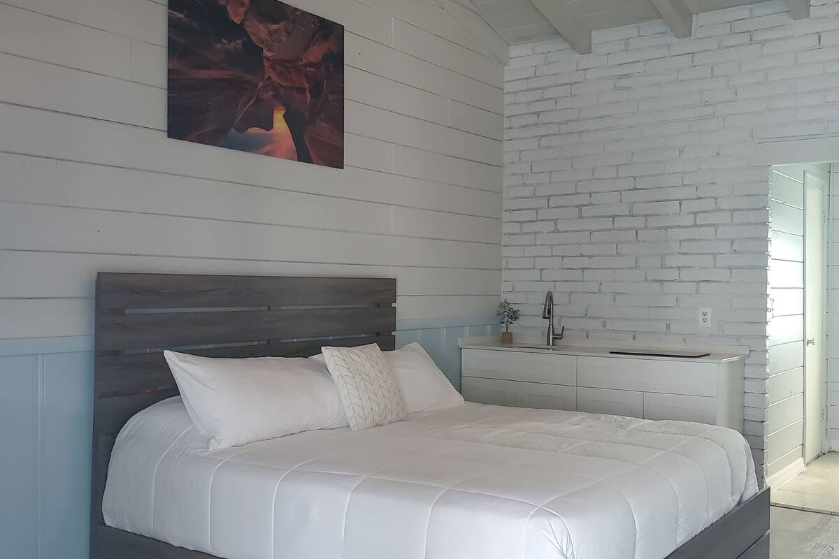 A bedroom with a wooden bed, white bedding, a white dresser, and a brick wall.