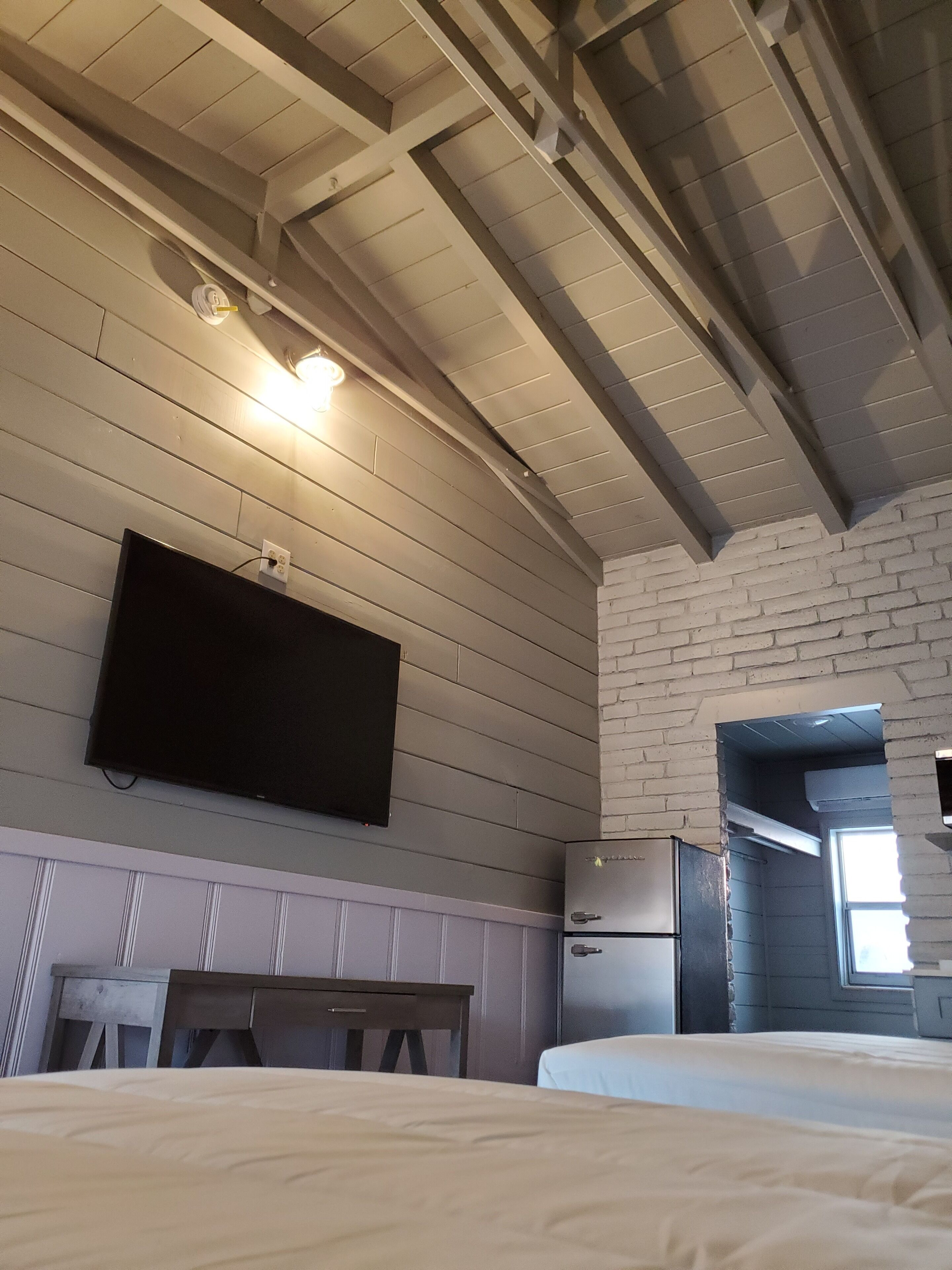 A room with a bed, a flat-screen TV mounted on the wall, a wooden bench, a brick wall, and a window.