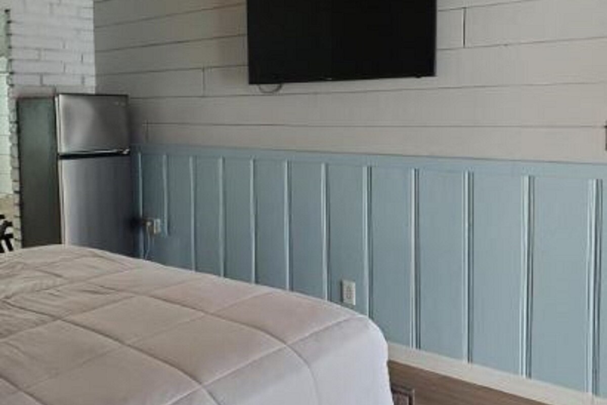 A bedroom with a bed, a flat-screen TV mounted on the wall, and a refrigerator in the corner.