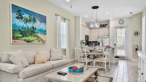 Un salon avec un canapé beige, un tableau représentant une scène de plage, un coin repas avec une table et des chaises blanches, et une cuisine avec des meubles blancs et une horloge au mur.