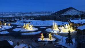 Paisaje nevado con una montaña al fondo, árboles iluminados y un camino que lleva a un edificio.