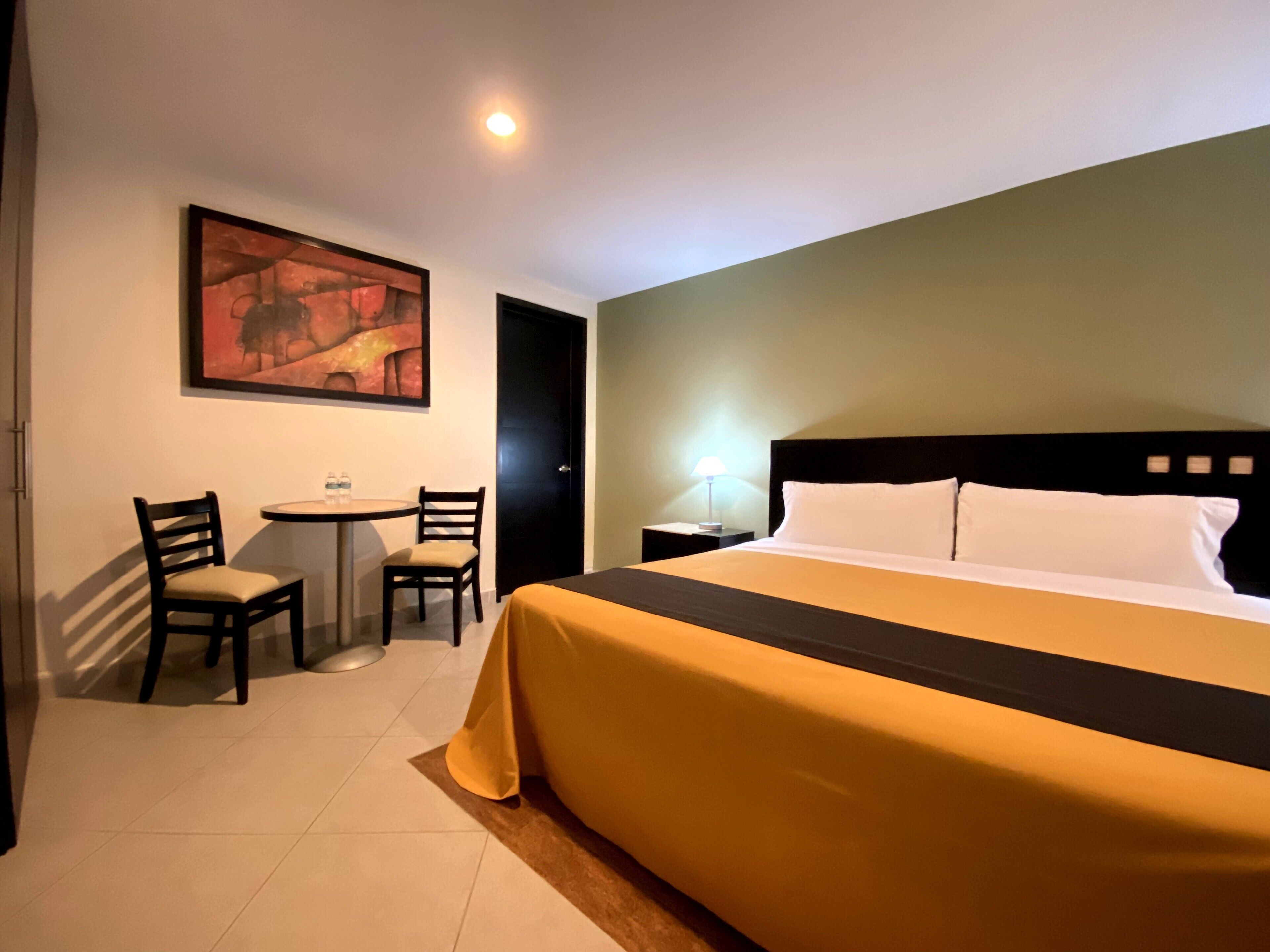 A hotel room with a bed, a desk, two chairs, a table, a painting on the wall, and a nightstand with a lamp.