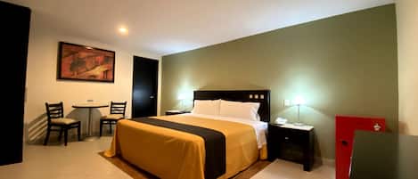 Habitación de hotel con cama, escritorio, dos sillas, un armario rojo y un cuadro en la pared.