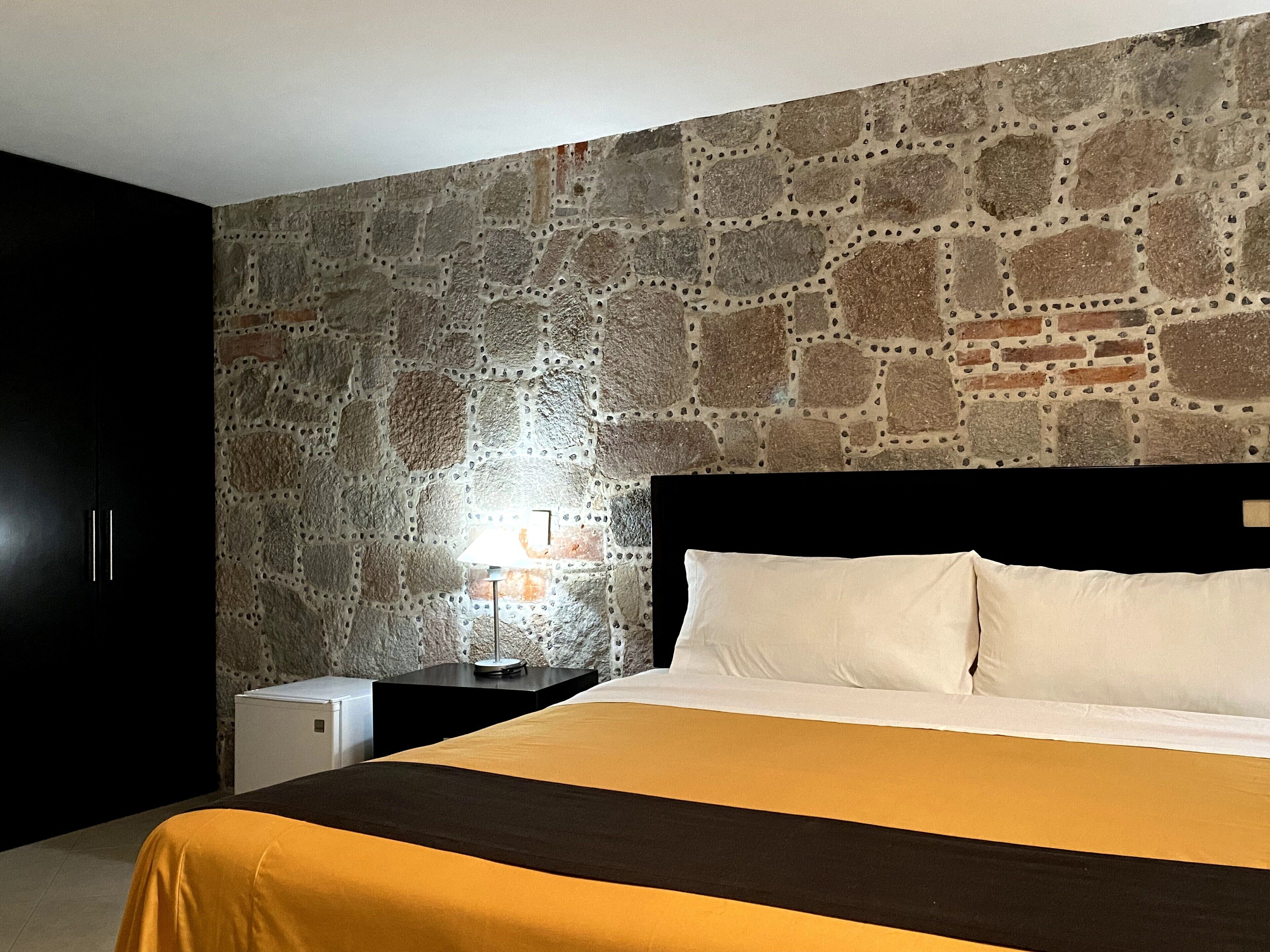 Un dormitorio con pared de piedra, una cama con ropa de cama blanca y amarilla, una mesita de noche con una lámpara y un electrodoméstico pequeño y blanco.