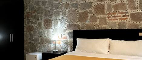 Un dormitorio con pared de piedra, una cama con ropa de cama blanca y amarilla, una mesita de noche con una lámpara y un electrodoméstico pequeño y blanco.