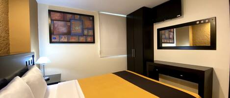 A hotel room with a bed, a desk, a chair, a lamp, a TV, and artwork on the wall.