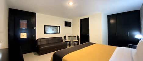 A hotel room with a bed, a sofa, a TV on the wall, and a desk with chairs.