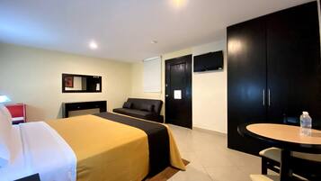 Habitación de hotel con cama, escritorio, silla, mesita, televisor y armario.