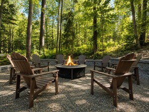 A circular fire pit surrounded by six Adirondack chairs in a forest setting.
