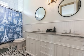Un baño con tocador blanco, dos lavamanos, inodoro y cortina de ducha con diseños parisinos.