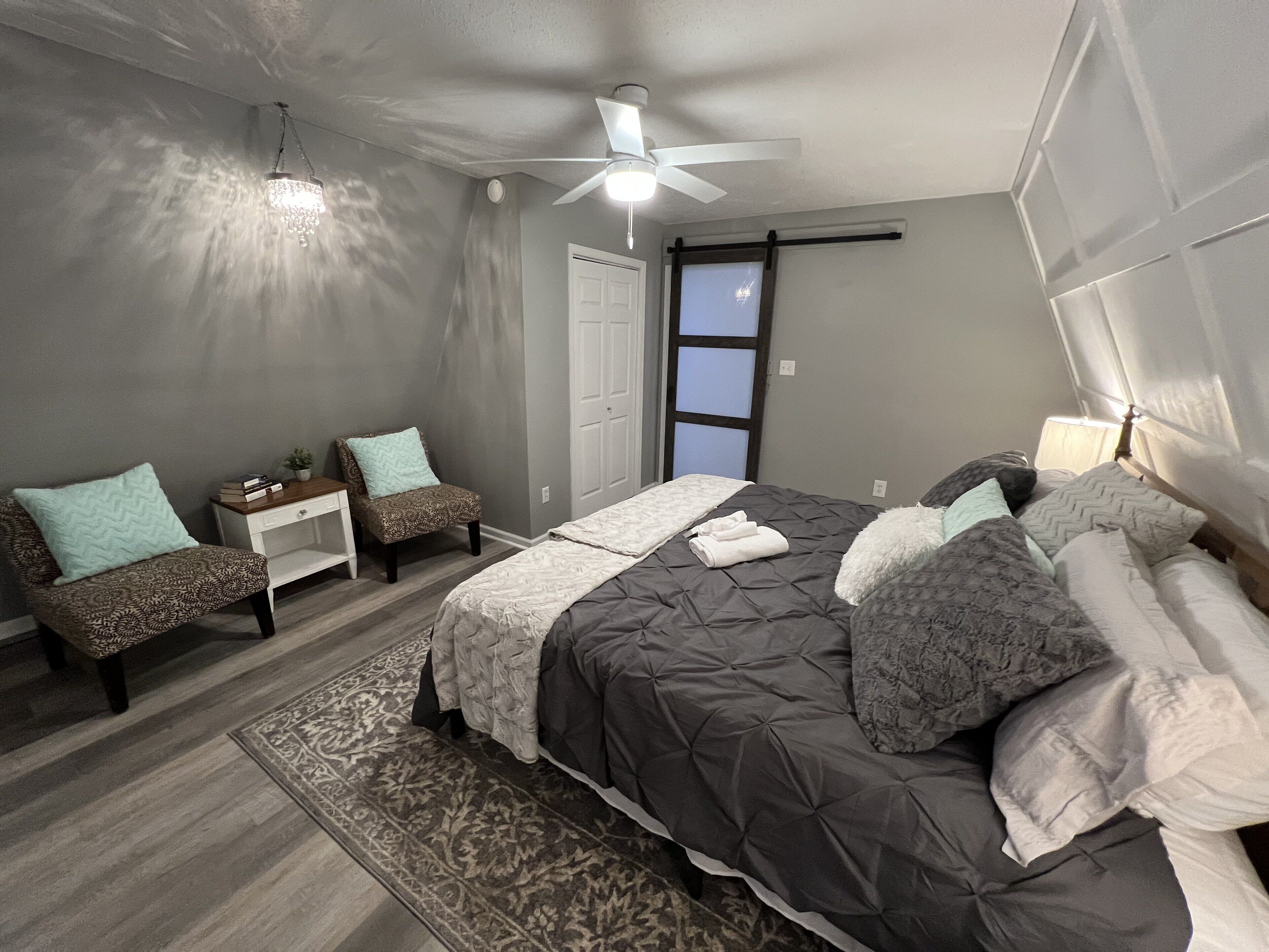 A bedroom with a bed, two chairs, a nightstand, a ceiling fan, and a window.