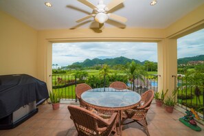 A balcony with a glass table and wicker chairs, overlooking a lush landscape with a pool and mountains.