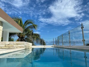 A modern pool with clear blue water, surrounded by stone walls and palm trees, under a partly cloudy sky.