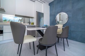 A modern kitchen with white cabinets, a large window, and a blue accent wall. There are two chairs and a table in the foreground. - Cayman Luxury Rentals at The Grove (Seven Mile Beach)