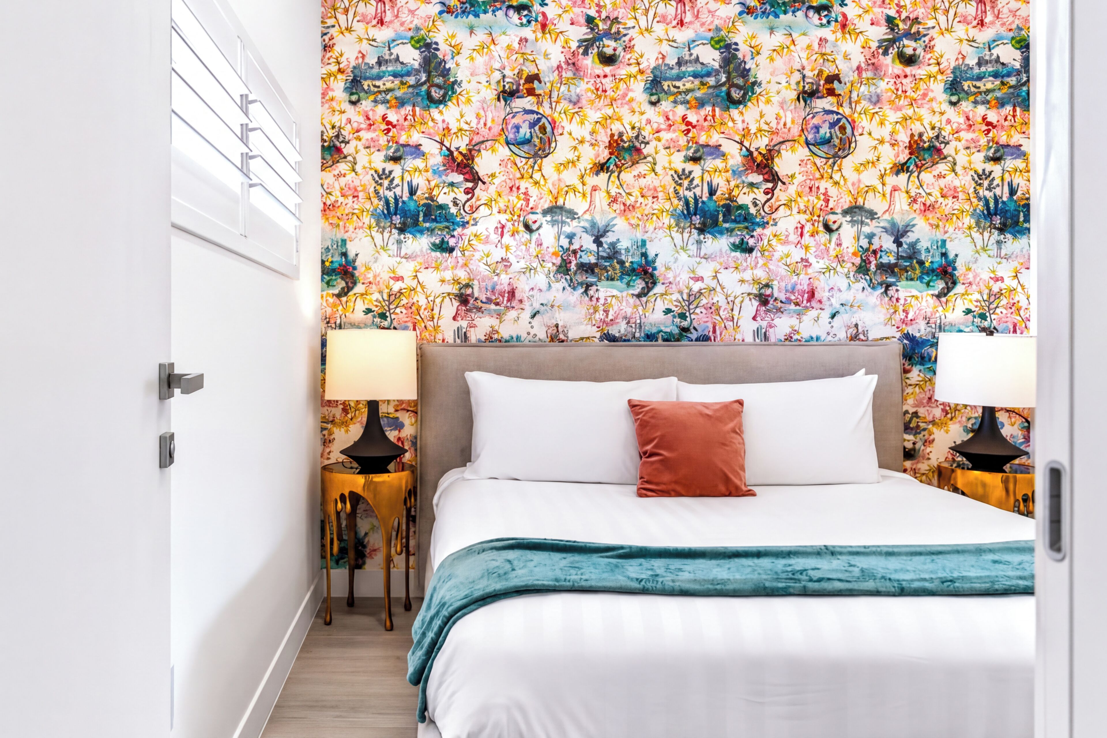 A bedroom with a floral wallpaper, a bed with white bedding and a teal throw, two bedside lamps, and a wooden floor.
