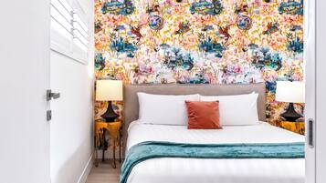 A bedroom with a floral wallpaper, a bed with white bedding and a teal throw, two bedside lamps, and a wooden floor.