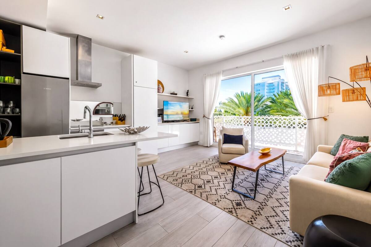 Una cocina moderna con una isla central, una zona de comedor con sofá y mesa, y un balcón con vistas a palmeras y edificios.