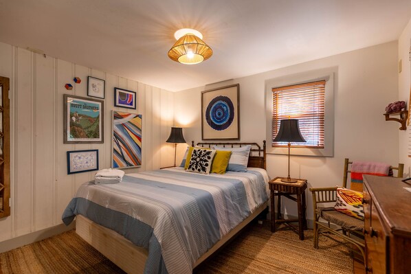 Un dormitorio ordenado con cama, mesita de noche, lámpara, silla y decoración en la pared.