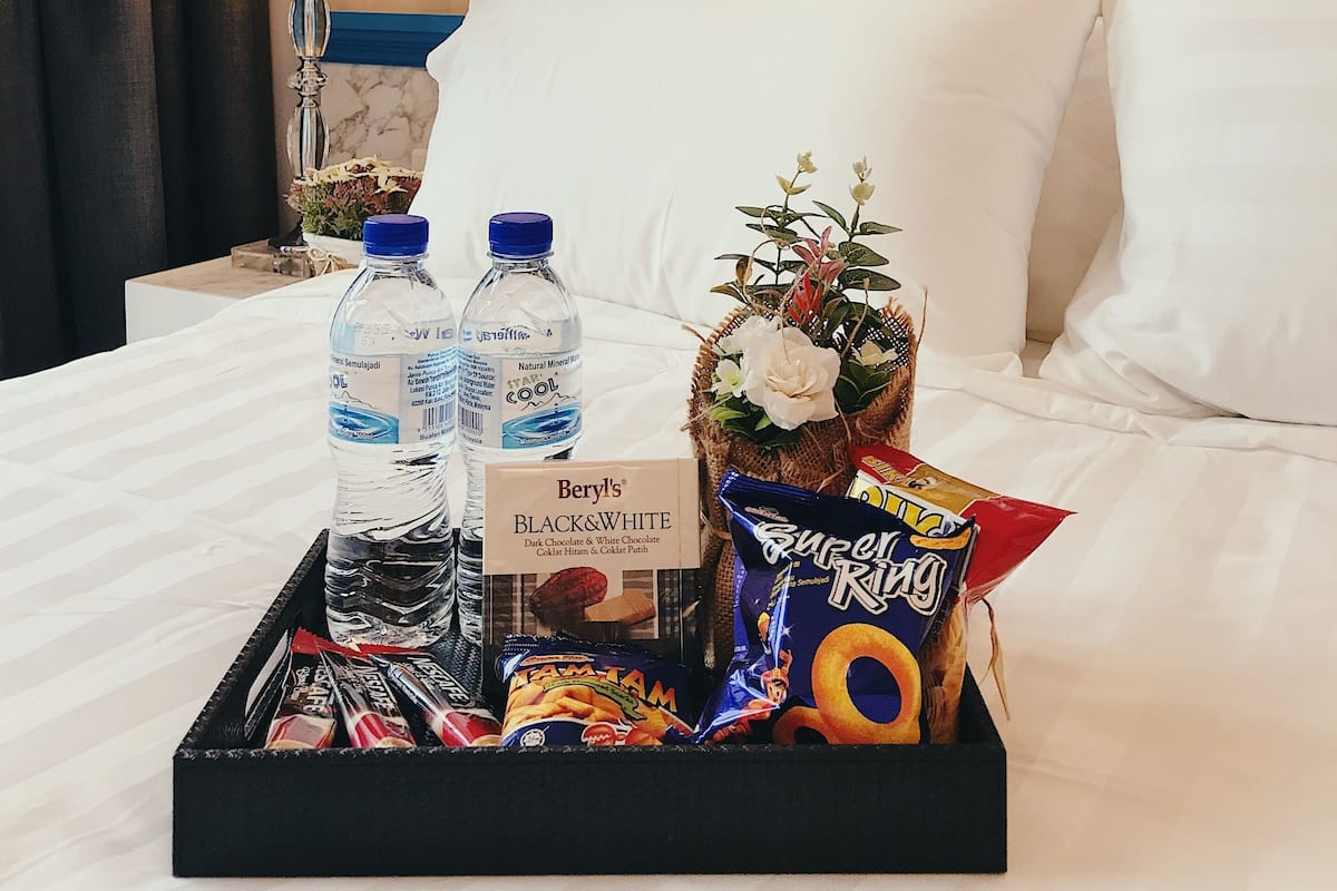 Habitación de hotel con cama, una bandeja de aperitivos, dos botellas de agua, una botella de chocolate Beryll's Black & White y una cesta de flores.