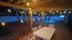 An outdoor patio with a wooden pergola, string lights, a white table and chairs, and a swimming pool in the background. - Coco's Beach House Residential Home w/ POOL (Bradenton)