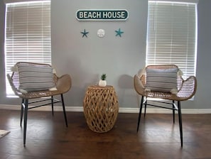 A room with two wicker chairs, a woven side table, and a wall sign reading "BEACH HOUSE" with star decorations.