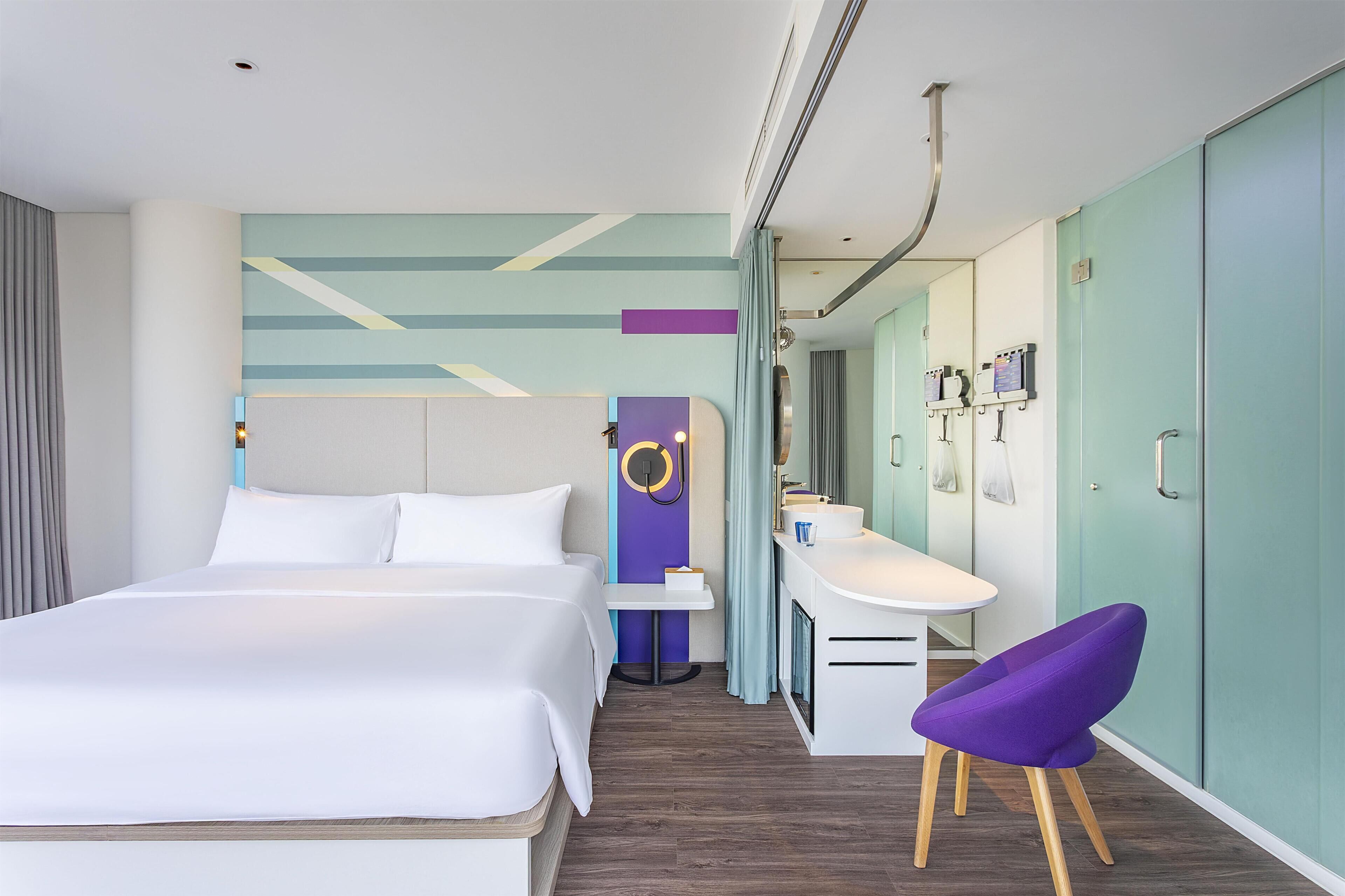 A modern hotel room with a large bed, a desk with a chair, a vanity, and a bathroom with a shower and toilet.