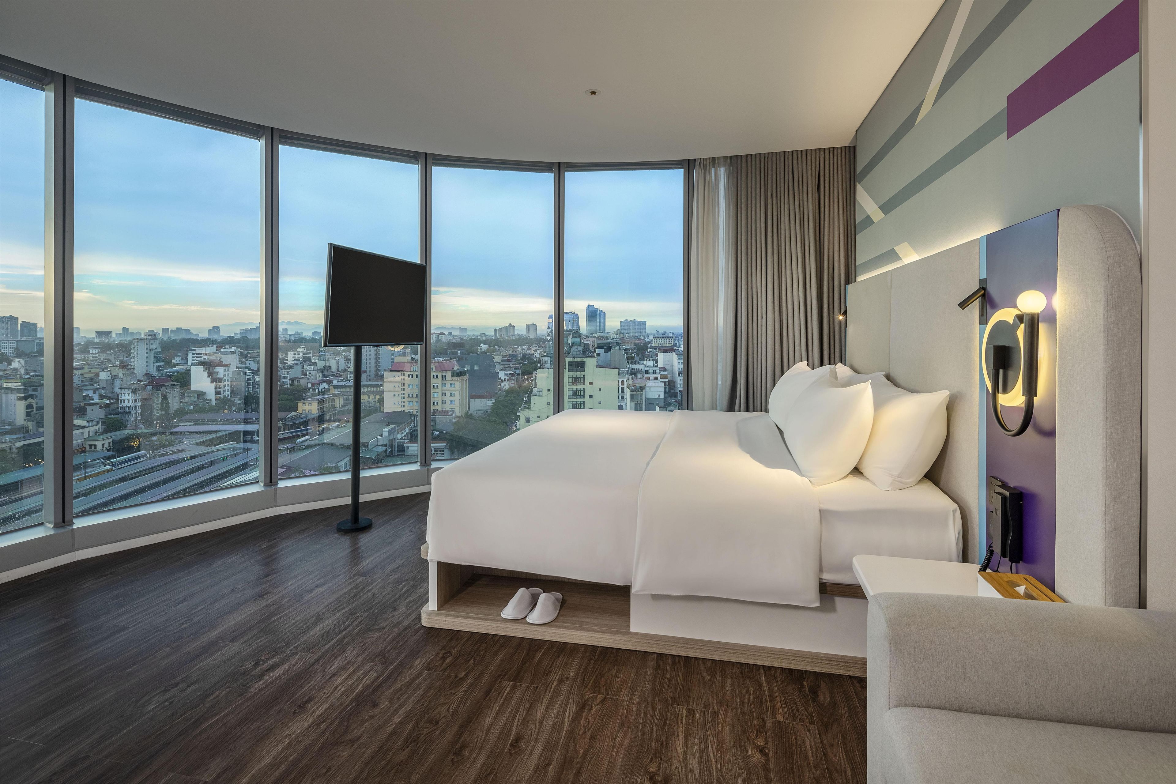 A modern hotel room with a large bed, a flat-screen TV, a city view, and a desk with a phone.
