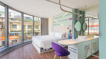 A modern hotel room with a large bed, a desk with a sink, and a view of the city through large windows.