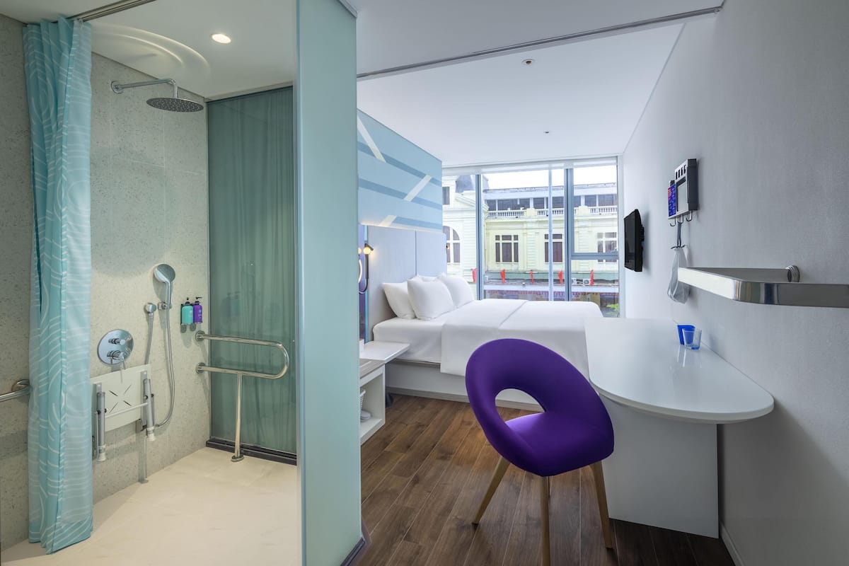 A modern hotel room with a shower, a bed, a small desk, and a window with a view of buildings.