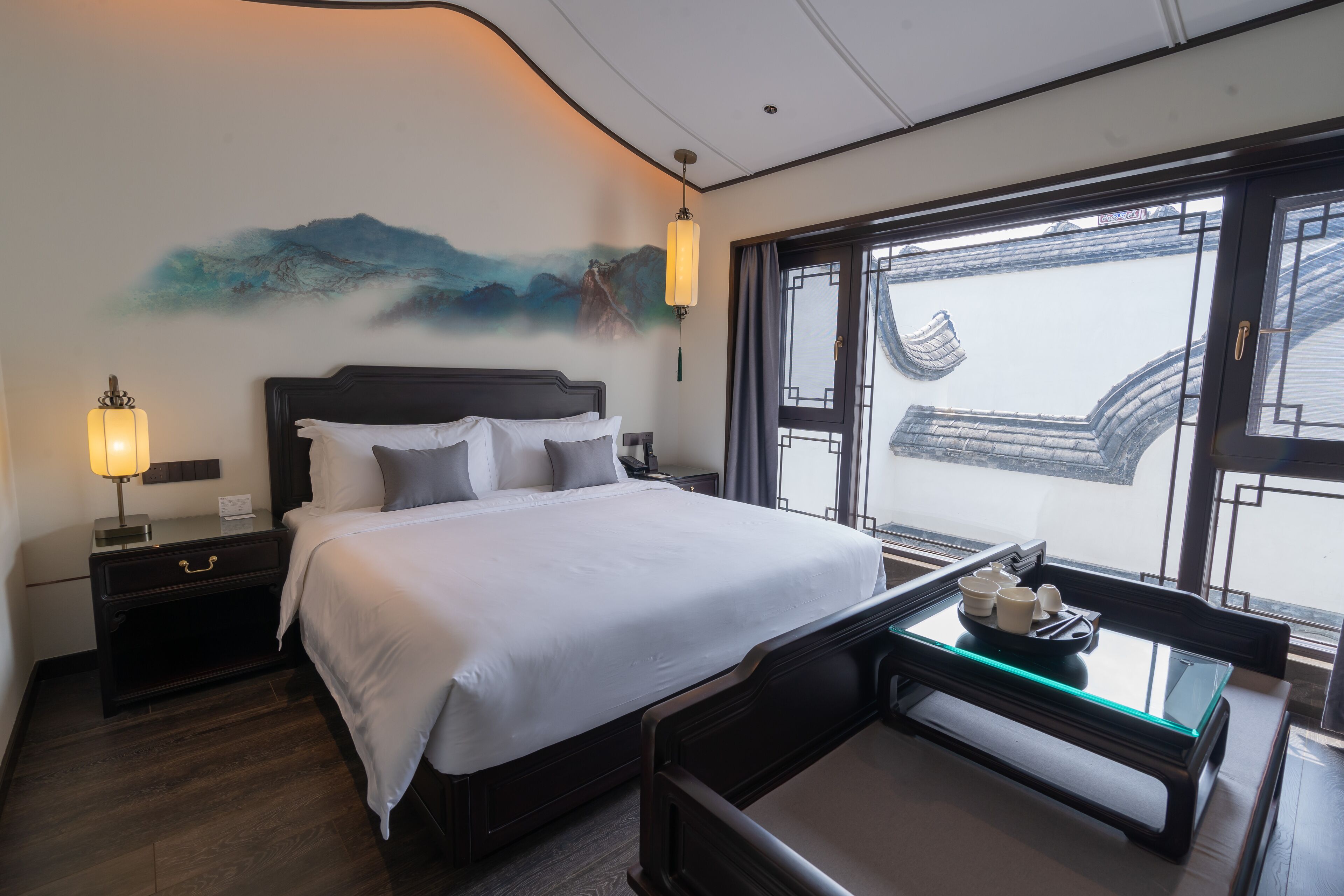 Habitación de hotel con una cama grande, una mesita de noche con lámpara, una pequeña mesa con servicio de té y una ventana con vistas a un edificio tradicional.