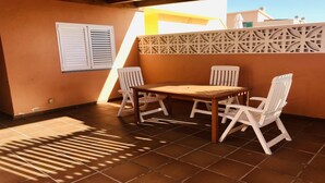 Un patio al aire libre con una mesa de madera y cuatro sillas blancas, una ventana con contraventanas y una pared decorativa.