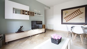 Ein modernes Wohnzimmer mit einem Flachbildfernseher, einer Holz-Unterhaltungsecke, einem Essbereich mit weißem Tisch und Stühlen sowie einem wandmontierten Regal mit der Aufschrift "DECO".