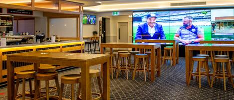 Zona de bar con taburetes y mesas de madera, una pantalla de televisión mostrando un evento deportivo y un cartel que indica el baño de hombres.