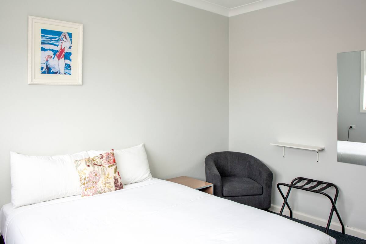 Une chambre d’hôtel comprenant un lit, une chaise, une table de chevet, une étagère fixée au mur et un tableau encadré accroché au mur.