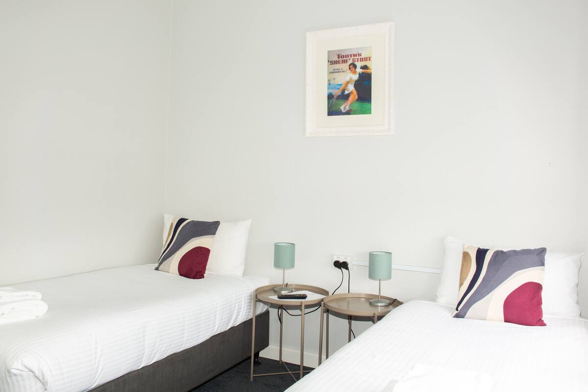Une chambre d’hôtel avec deux lits, chacun agrémenté de draps blancs et d’un oreiller rouge. On y trouve deux tables de chevet avec des lampes, ainsi qu’un tableau encadré accroché au mur.
